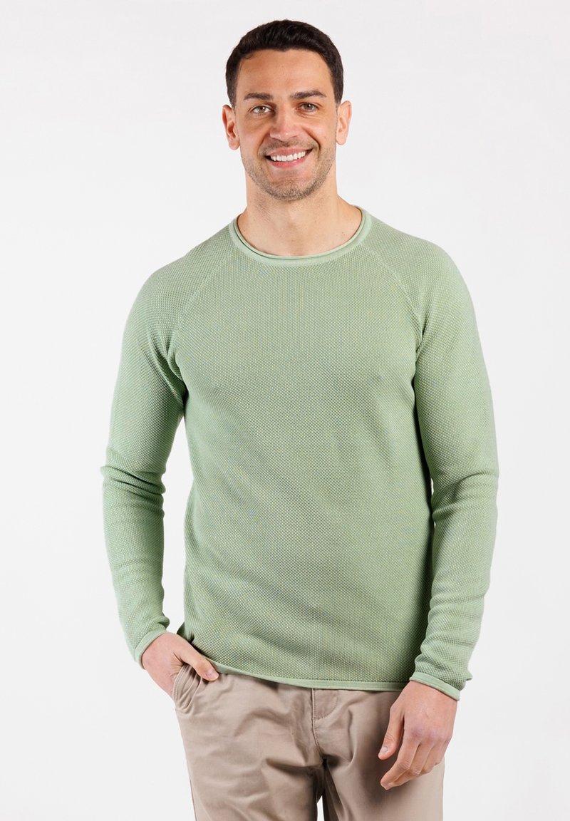 Un uomo sorridente sta in piedi con una mano in tasca, indossando un maglione tessuto verde chiaro e pantaloni beige su uno sfondo bianco.