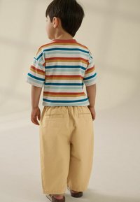 Gestreept T-shirt met korte mouwen in wit, blauw, rood en geel; loszittende beige broek met achterzakken, gedragen met beige sandalen.