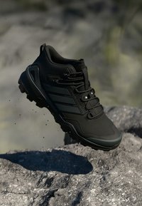 Schwarzer Wanderschuh mit dicker Sohle und robustem Profil in der Luft über felsigem Gelände, der kleine Staubpartikel aufwirbelt.