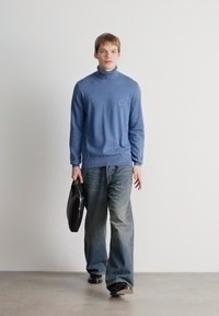 Suéter azul de gola alta com tecido texturizado, combinado com jeans de corte largo de cor azul clara. Modelo segura uma pasta preta, em pé contra uma parede branca.