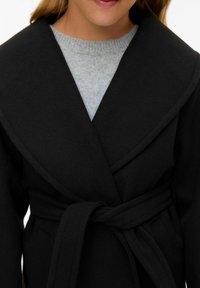 Cappotto nero avvolgente con ampio colletto e legatura in vita. Realizzato in tessuto morbido, presenta maniche lunghe e tasche applicate. Maglione grigio chiaro sotto.