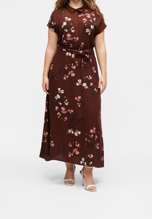 Kvinde iført en brun blomsterprintet maxikjole med bånd i taljen og korte ærmer, kombineret med lyse beige sandaler med høje hæle.