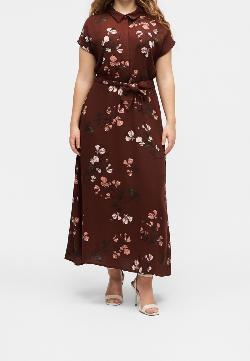 Femme portant une robe maxi marron à fleurs avec une ceinture nouée à la taille et des manches courtes, associée à des sandales à talons hauts beige clair.