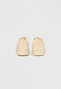 Beige treningssko med teksturert overdel, perforerte detaljer, polstret krage og kontrasterende stilisert logo på siden.