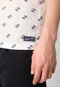 Short-sleeve shirt in light beige fabric with navy pattern. Features a logo patch and a relaxed fit, paired with dark jeans.