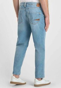 Jeans de mezclilla azul claro con corte recto, que cuentan con bolsillos traseros y una etiqueta de cuero marrón en la cintura. Combinados con zapatillas blancas.