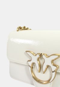Borsa a tracolla in pelle verniciata bianca con una catena dorata e una chiusura a forma di farfalla con un anello dorato circolare. Logo "PINKO" visibile.