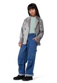 Blazer plateado con lentejuelas, de doble botonadura, camiseta verde y jeans cargo azules sueltos con bolsillos laterales, combinados con zapatos negros.