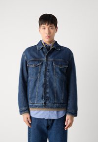 Jeansjacka i mörkblått med knäppning fram, två bröstfickor och en kontrasterande beige krage. Bärs över en ljusblå skjorta och jeans.