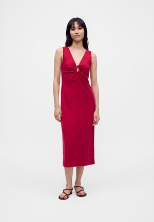 Femme debout portant une robe midi rouge sans manches avec une ouverture en forme de goutte devant et un détail de lien, associée à des sandales à lanières foncées sur un fond uni.