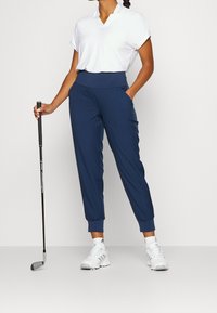 Marinblå byxor med uppvikta avslut och fickor, i kombination med en vit skjorta med krage. Modellen håller en golfklubba, iklädd vita sneakers med detaljer.