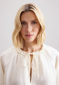 Blusa morbida color crema con un collo a balze arricciato e chiusura con lacci, realizzata in tessuto testurizzato, con una sottile apertura plissettata.