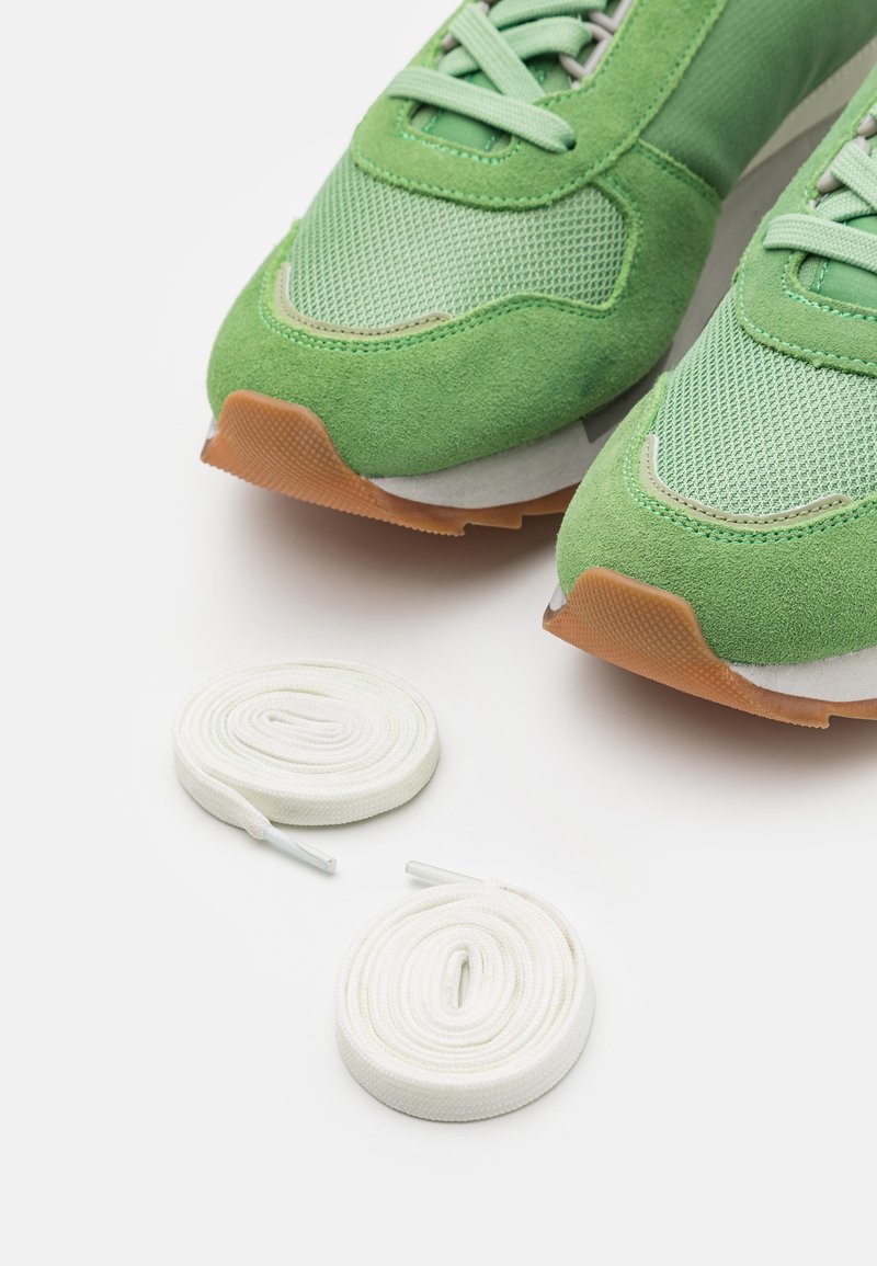 Zapatillas deportivas verdes fabricadas en malla y gamuza, con suela de goma y cordones blancos. Dos cordones blancos enrollados están colocados al lado de las zapatillas.