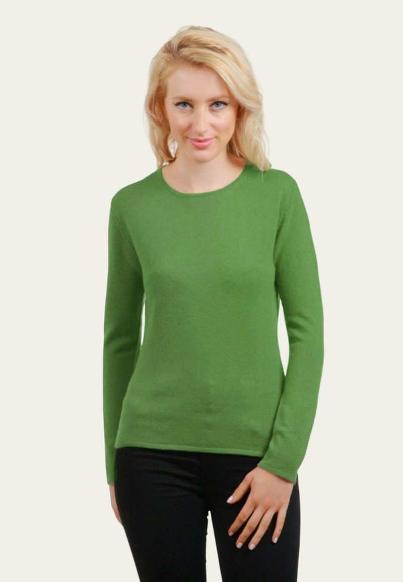 Femme blonde à cheveux ondulés portant un pull vert à manches longues et un pantalon noir, se tenant avec une main reposant sur sa hanche.