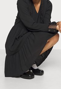 Robe noire à manches longues avec un ourlet en couches et volants. Présente des détails en dentelle sur les manches et de grands boutons. Portée avec des chaussures à plateforme noires.