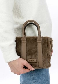 Borsa in finta pelliccia marrone con manici in pelle liscia, forma rettangolare e un piccolo etichetta con logo, appoggiata su un maglione bianco.