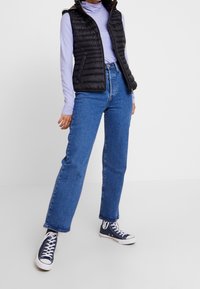 Czarna pikowana kamizelka z przodu na zamek, bez rękawów, na jasnofioletowej bluzce z długim rękawem, do tego proste niebieskie dżinsy i granatowe trampki.