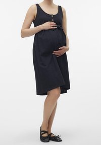 Gravidklänning i marinblått, ärmlös design, med framsidans knappar och knytning i midjan, tillverkad av mjukt och lätt tyg. Bärs med svarta platta skor.
