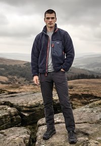 Ung mann står på steinete terreng iført en marineblå fleecejakke og mørke bukser med et skyet, kupert landskap i bakgrunnen.
