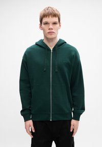 Mørkegrøn zip-up hoodie med en frontlomme, snørelukning i hætten og et diskret logo på brystet. Lavet af blødt, tekstureret stof.