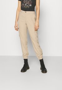 Beige Cargo-Hosen mit elastischen Bündchen, ausgestattet mit seitlichen Taschen und einem Knopfverschluss. Kombiniert mit schwarzen Schnürstiefeln und einem Grafik-T-Shirt.