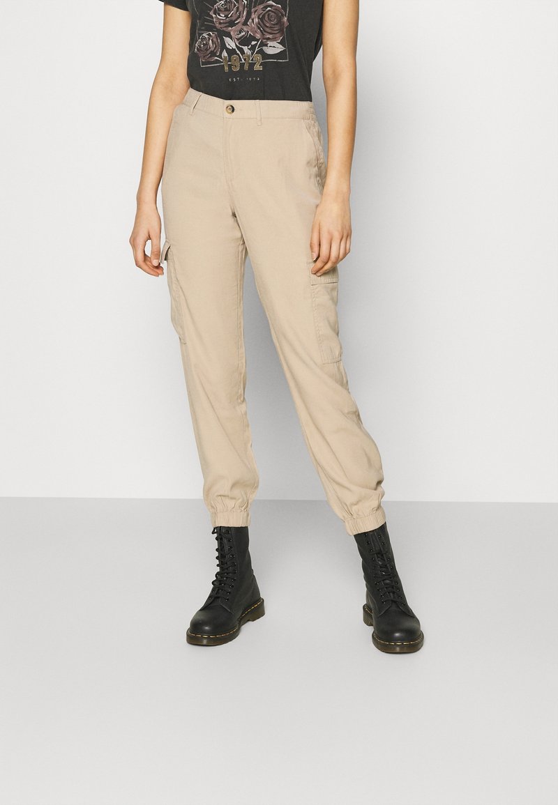 Beige Cargo-Hosen mit elastischen Bündchen, ausgestattet mit seitlichen Taschen und einem Knopfverschluss. Kombiniert mit schwarzen Schnürstiefeln und einem Grafik-T-Shirt.