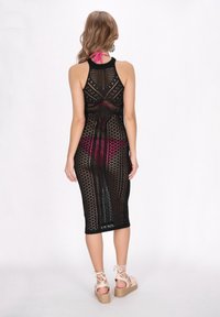 Vestido negro de punto con diseño de rejilla abierta, con cuello halter y corte a la altura de la rodilla, que revela un color rosa debajo. Llevado con sandalias claras.