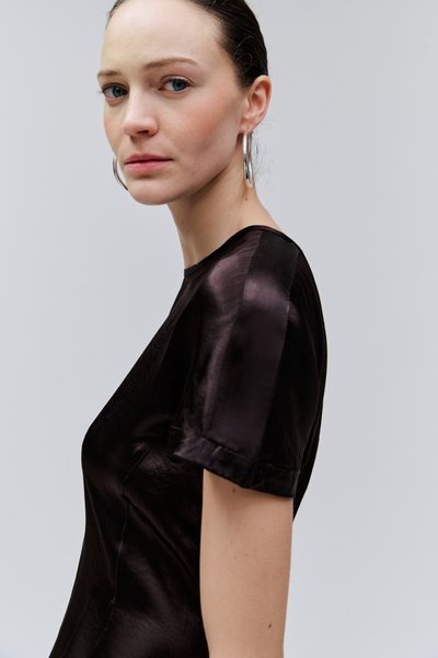 Femme aux yeux bleus et à la peau claire portant un haut court noir brillant à manches courtes et de grandes boucles d'oreilles argentées en forme d'anneau, regardant légèrement sur le côté.