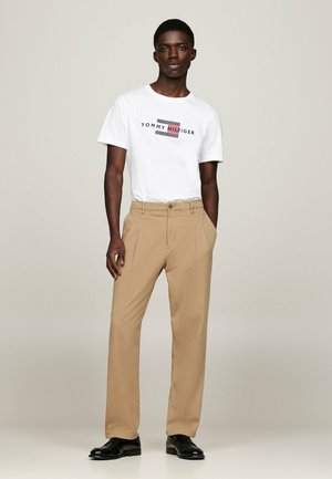 T-shirt blanche avec un logo rouge et bleu, accompagnée d'un pantalon beige. Le pantalon est droit avec une texture lisse et une fermeture à bouton. Chaussures noires.