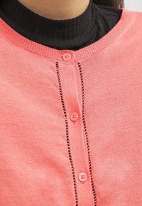 Cardigan i ljusrosa stickad med rund halsringning, med fyra rosa knappar och dekorativa stygn längs knapppkanalen.