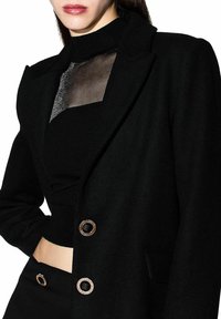 Blazer negro con cuello con solapa, panel transparente en el pecho y botones circulares metálicos. Presenta un ajuste entallado y una textura suave.