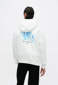 Biela mikina s kapucňou a veľkým modrým grafickým prvkom na chrbte, ktorý obsahuje abstraktné línie a logo Nike. Mäkkučká látka s voľným strihom.