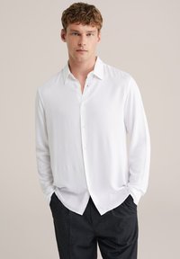 Camicia bianca a maniche lunghe con bottoni, in tessuto leggero, taglio rilassato, con colletto a punta e tessuto semplice. Abbinata a pantaloni scuri.