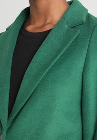 Blazer de lana verde con solapa de muesca, acabado texturizado y corte entallado, combinado con una camisa negra.