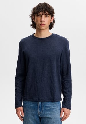 Młody mężczyzna o kręconych włosach, ubrany w granatową bluzę z długim rękawem i niebieskie dżinsy, stojący na tle jednolitego jasnego tła.