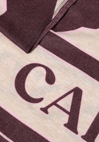 Tessuto a strisce marrone e rosa chiaro con una texture morbida, caratterizzato da lettere in grassetto e un dettaglio di cucitura sul bordo.