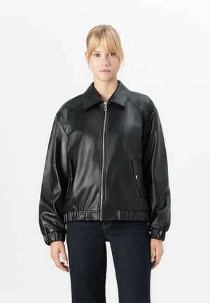 Femme aux cheveux blonds portant une veste bomber en similibois noir avec des poignets élastiques et un jean foncé, se tenant contre un fond blanc.