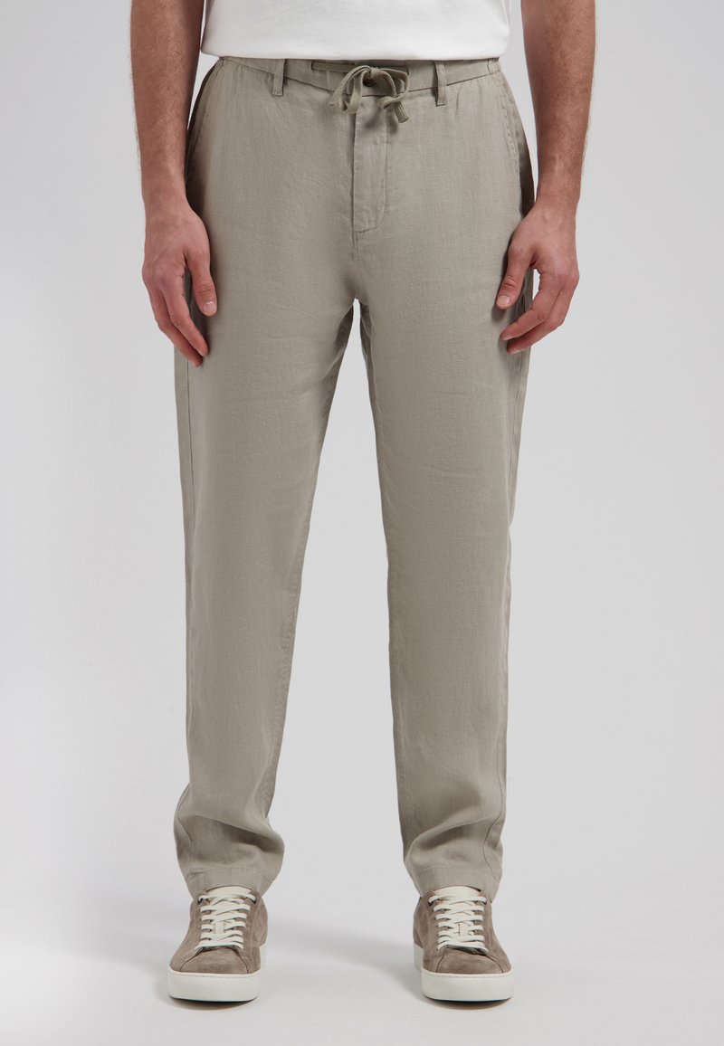 Homme portant un pantalon à cordon de serrage beige clair et des baskets beiges, debout devant un fond uni.