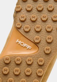Gros plan sur la semelle marron texturée d'une chaussure, avec des crampons hexagonaux embossés d'un "H" et le nom de la marque "HOFF" au centre.