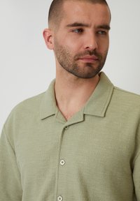 Hellgrünes, strukturiertes Button-Down-Hemd mit kurzem Kragen und drei Knöpfen, das ein dezentes Gewebemuster aufweist und eine lässige Passform hat.