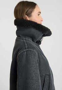 Giacca in denim nera con un colletto alto e morbido. Presenta un tessuto testurizzato, cuciture visibili e una chiusura sicura al collo.