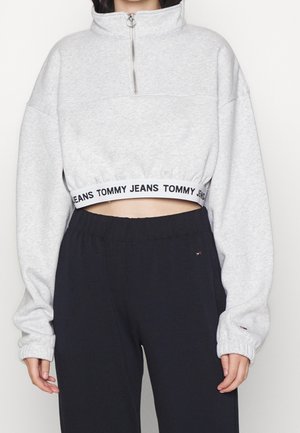 Personne portant un sweat-shirt court gris clair avec une demi-fermeture éclair et un bandeau élastique portant la mention "Tommy Jeans", associé à un pantalon noir.