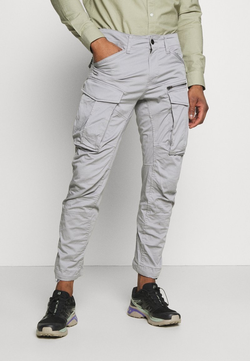 Light grey cargo pants with multiple pockets, button closure, and a tapered leg design, made of lightweight fabric. Worn with black sneakers.