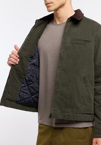 Olive green jacket with a corduroy collar, zip closure, and a quilted dark inner lining. Features a chest pocket and side zipper pockets.