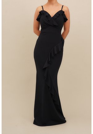 Kobieta ubrana w długą, czarną, bezrękawnikową sukienkę z falbankami na dekolcie i rozciągającymi się w dół spódnicy.