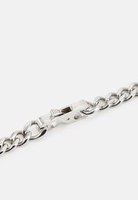 Bracelet en chaîne en métal argenté avec de grands maillons et un fermoir, gravé de "GUESS EST. 1981." Texture lisse et polie.