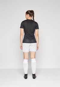 Schwarzes Sportshirt mit weißen Seiteneinsätzen, kombiniert mit hellgrauen Shorts und weißen Kniestrümpfen. Schwarze Sportschuhe runden den Look ab.