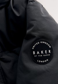 Svart tygjacka med en cirkulär märkespatch med texten: "BAKER by TED BAKER, UNITED KINGDOM, LONDON." Slät textur, minimalistisk design.