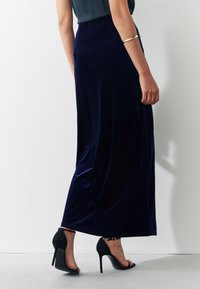 Donna che indossa una lunga gonna in velluto blu navy e sandali neri con tacco alto, in piedi davanti a una parete bianca uniforme.