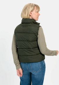 Dunkelgrüwestes Pufferweste mit hohem Kragen und horizontalem Steppmuster. Getragen über einem langen beigen Oberteil, kombiniert mit blauen Jeans.